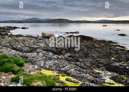 Rocky shore of Isle of Iona with sailboats on Sound of Iona and view of Fionnphort Isle of Mull mountains Inner Hebrides Scotland UK Stock Photo