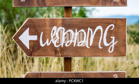 Handmade wedding sign pointing left towards the ceremony, white paint on brown base with blurred country background, outdoors Stock Photo