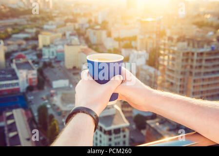 Close up man's hands holding take away paper cup with morning hot drink - coffee or tea with inspiring view on abstract city background at sunrise or sunset. Soft selective focus. Copy space. Stock Photo