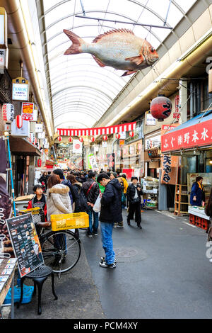 Kuromon Ichiba, Osaka's kitchen food market. View along arcade with shoppers and tourists walking through. Model fish hanging from roof. Wintertime. Stock Photo