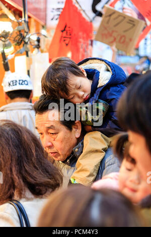 Japanese young child, boy, asleep on his father back as he is carried through a busy market in the day time. Candid shot at Kuromon Ichiba, Osaka. Stock Photo