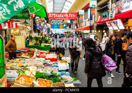 Kuromon Ichiba, Osaka's kitchen food market. View along arcade with various stalls, foreground green grocers. Busy, many people. Winter. Stock Photo