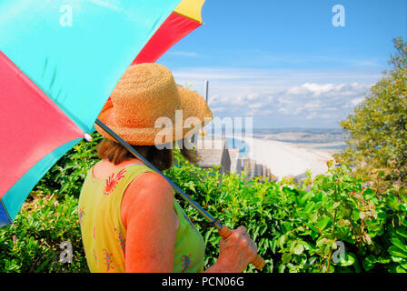 Portland. 3rd August 2018. A woman with a rainbow sunshade looks out over Chesil Beach, on a perfect summer's day in Fortuneswell Credit: stuart fretwell/Alamy Live News Stock Photo