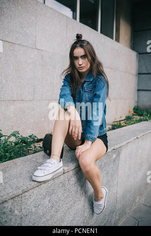 Teen girl posing in city wearing casual outfit: shorts, blue jeans jacket and white sneakers. Portrait of female with trendy bun hairstyle sitting nea Stock Photo