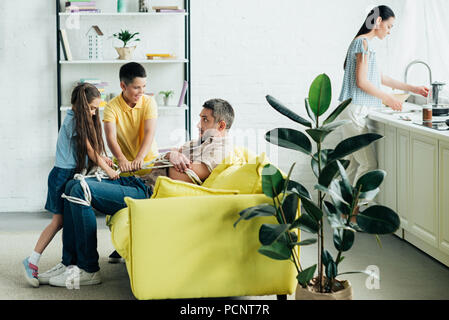children tying father with rope at home, mother washing dishes Stock Photo