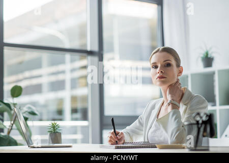 beautiful young businesswoman sitting at workplace in modern office and looking at camera Stock Photo