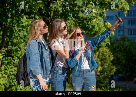 Three girl friends girl schoolgirl. Summer in nature. In jeans clothes behind backpacks. Take pictures of yourself on smartphone, sunglasses. Sends an air kiss. Stock Photo