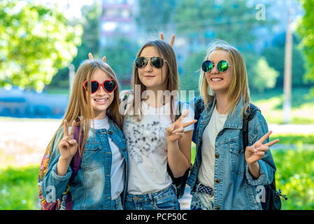 Three girl friends girl schoolgirl. Summer in nature. In jeans clothes behind backpacks. In sunglasses. Gesture of hands shows Hello, Victory. Stock Photo