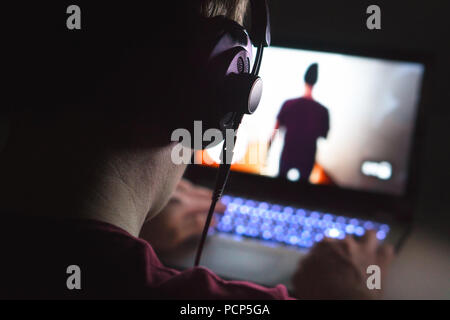 Playing video games with laptop. Young man plays action game on computer. Back view of gamer with headphones in dark or late at night. Stock Photo