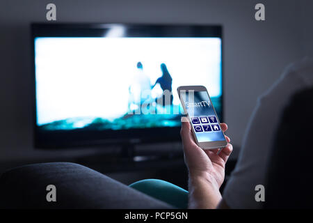 Man watching television and using smart tv remote control application on mobile phone. Choosing movie stream, switching channel or changing settings. Stock Photo