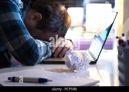 Sad and tired man in home or modern business office at night. Unemployed job seeker or businessman with ripped paper and laptop on table. Stock Photo
