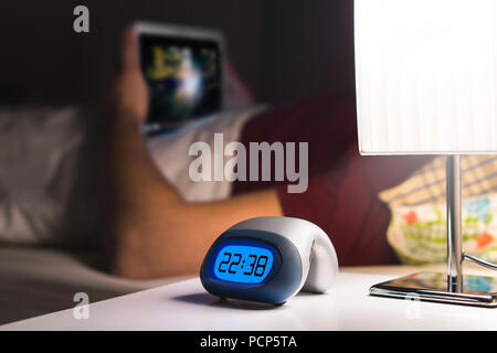 Man using tablet or watching movie or series stream online in bed before falling asleep. Digital alarm clock on nightstand in bedroom. Stock Photo