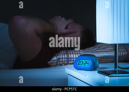 Insomnia and sleepless concept. Man unable to sleep. Exhausted and tired. Covering face with hand. Alarm clock on nightstand and bed in bedroom. Stock Photo