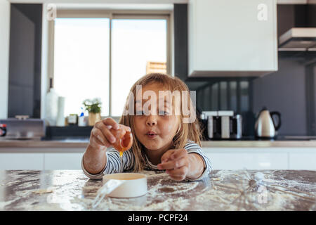 Cute little girl breaking an egg into a small pan over kitchen counter. Beautiful young girl baking in the kitchen. Stock Photo
