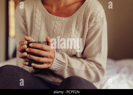 Close up of woman wearing sweater holding a cup of coffee while sitting on bed. Cropped shot of woman having morning coffee in bed. Focus on hands hol