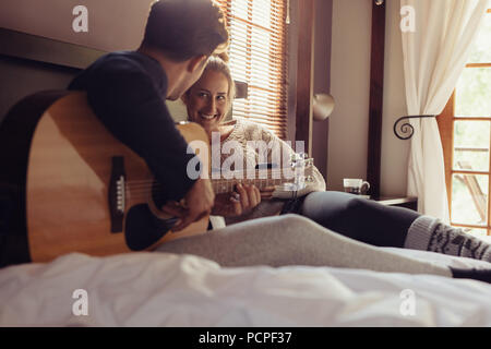 Young man playing guitar for his girlfriend in bed. Loving couple resting in hotel room and playing guitar in bed. Stock Photo