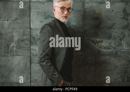 Portrait of confident mature businessman standing against grey background. Handsome male standing with his hands in pockets and looking at camera. Stock Photo