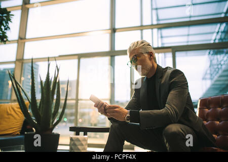 Mature businessman using mobile phone while sitting in office lounge. Senior executive texting on cell phone in office lobby. Stock Photo