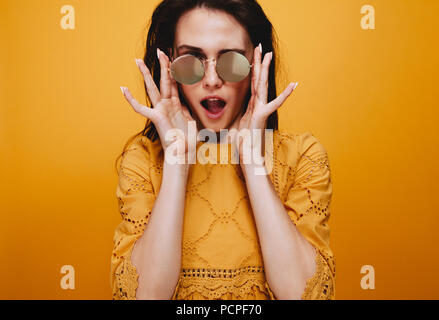 Portrait of beautiful female model wearing funky sunglasses. Attractive young woman in orange dress looking at camera with mouth open. Stock Photo