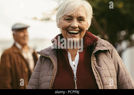 Portrait of cheerful senior woman in warm clothes looking at camera and laughing with a man in background. Old woman outdoors on a winter day. Stock Photo