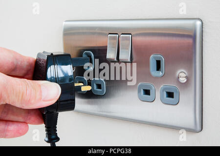 A person's hand holding an unplugged three pin plug and plugging it in to an electric wall socket switched off. England, UK, Britain