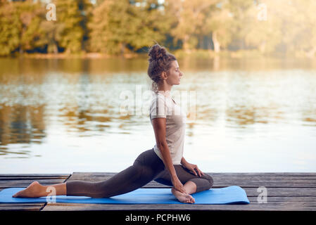 Attractive young woman working out on mat on a wooden deck above a lake or river doing a yoga pose with her eyes closed and a serene expression Stock Photo