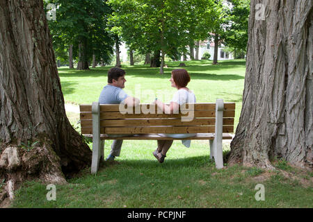 A man and a woman sit together on a park bench surrounded by huge trees, looking at one another and smiling Stock Photo