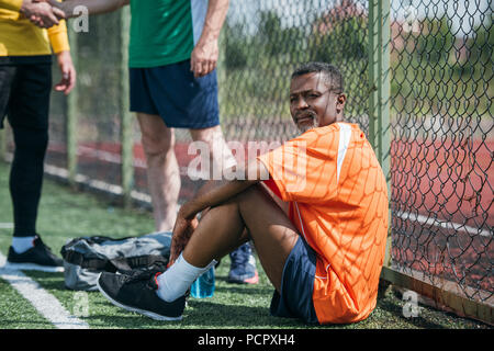 selective focus of elderly african american man resting on football field while friends shaking hands after match Stock Photo