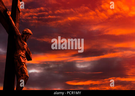 Jesus on a cross against burning red sunset sky with clouds and little patches of light coming through Stock Photo