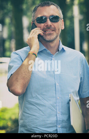 Businessman holding his laptop using his smartphone, standing on the street Stock Photo