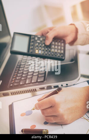 Womans hand holding calculator and writing with pen, financial chart report and notebook on desk at office. Stock Photo
