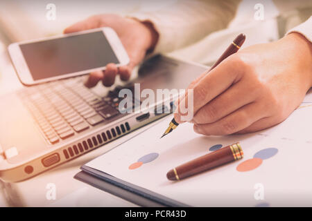 Womans hand holding smart phone and writing with pen, financial chart report and notebook on desk at home office. Stock Photo