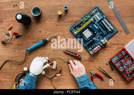 cropped image of man with prosthetic arm holding wires and screwdriver surrounded by instruments on wooden table Stock Photo