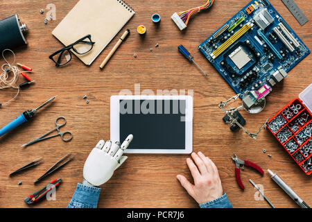 cropped image of man with robotic hand using digital tablet at table surrounded by tools Stock Photo