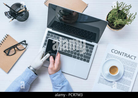 cropped image of businessman with prosthetic arm checking smartphone at table with laptop in office Stock Photo