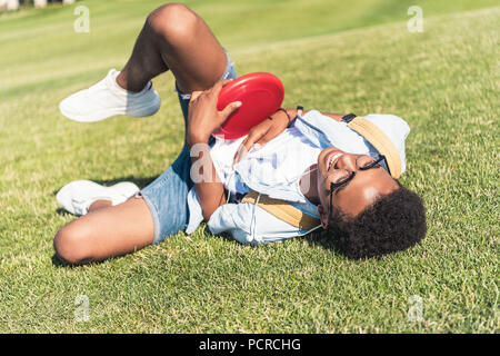 happy african american teenager holding lying disk and lying on grass in park Stock Photo