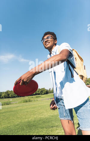 smiling african american teenager with backpack throwing flying disc in park Stock Photo