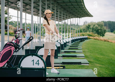 pensive female golfer in polo and cap with golf club looking away at golf course Stock Photo