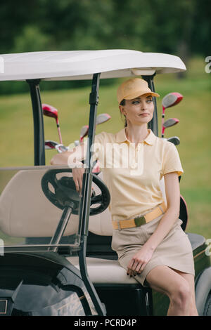 portrait of smiling woman in polo and cap sitting on golf cart and looking away Stock Photo