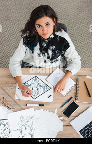 overhead view of female artist painting and looking at camera while sitting at table with laptop and smartphone