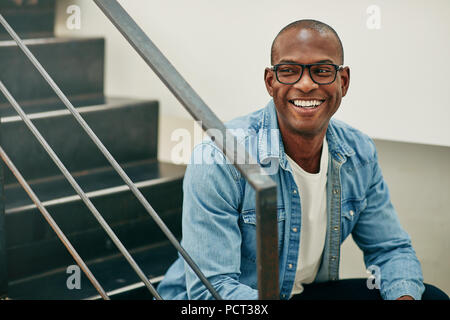 Confident young African businessman wearing glasses and laughing while sitting alone on stairs in a modern office Stock Photo