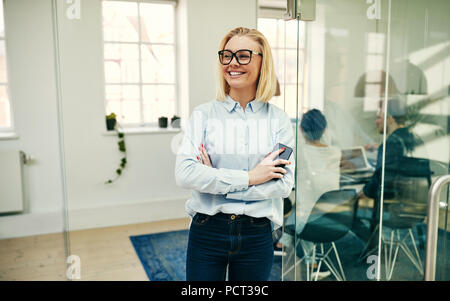 Smiling young businesswoman wearing glasses and leaning against a glass wall in an office holding her cellphone Stock Photo