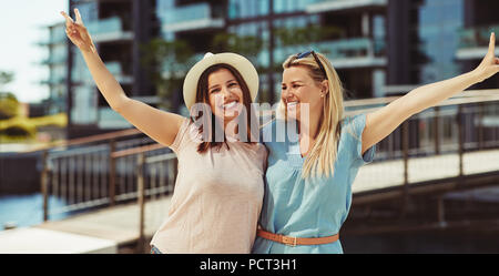 Two laughing young female friends standing arm in arm together in the city on a  summer day having a good time Stock Photo