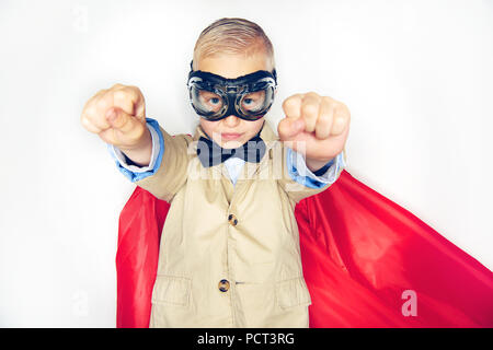 Cute little boy superhero in a suit and bowtie wearing a red cape and goggles pretending to fly while standing against a white background Stock Photo