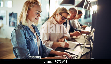 Smiling young female designer working on a laptop while sitting in a row with colleagues at a table in an office Stock Photo