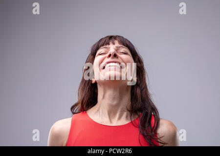 Elated relaxed woman with a wide warm beaming smile tilting her head back with closed eyes isolated on grey Stock Photo