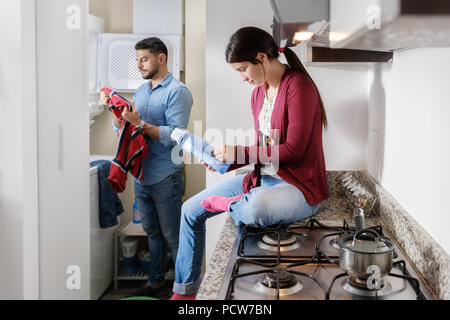 Young couple doing housework and chores. Man putting clothes inside washing machine and girlfriend sitting on kitchen counter, reading label on cleans Stock Photo