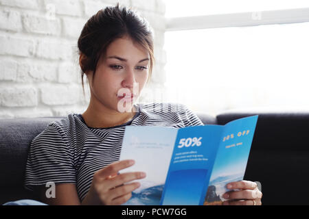 Young hispanic woman laying on couch at home, holding a travel flyer and planning next trip. Copy space on window. Stock Photo