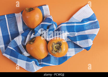 Three ripe persimmons on checkered blue towel on an orange background. Concept of autumn seasonal fruits, flat lay top view Stock Photo