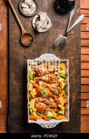 Top view of Chicken drumsticks with vegetables in baking casserole on rustic wooden background with cooking spoon. Simple country food concept Stock Photo